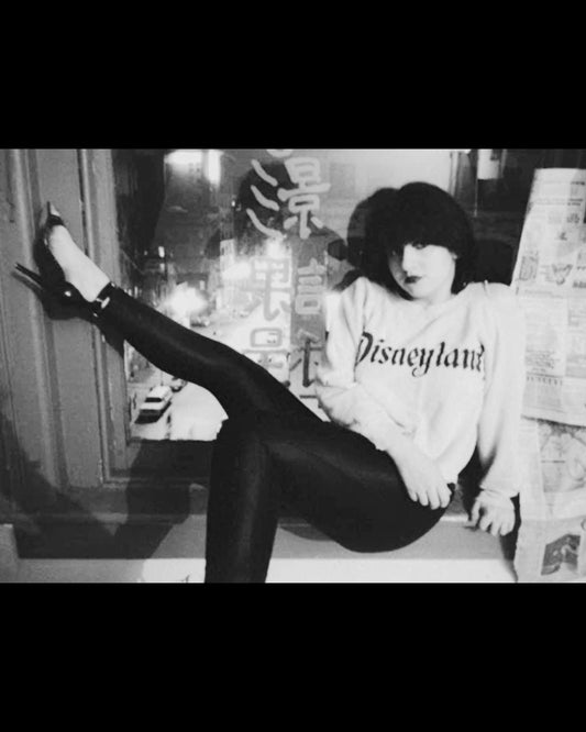 Lydia Lunch sitting in a Delancey Street loft in 1977/78, wearing a Disneyland sweatshirt and black trousers, photographed by GODLIS.